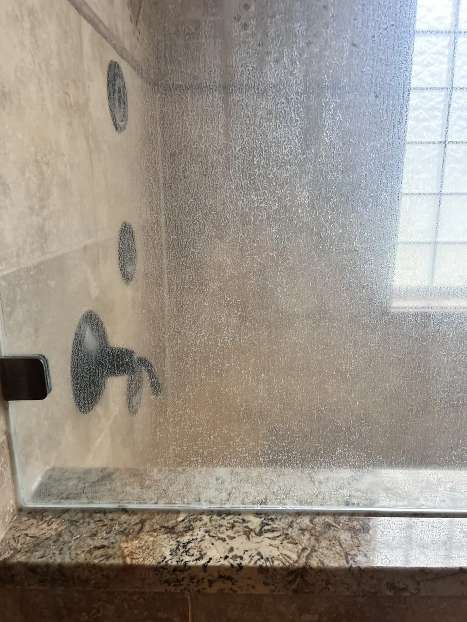 Cloudy residue on shower glass obscuring the tan tile and glass-block window
